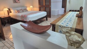 Une chambre d’hôtel comprenant un lit, une table de chevet, une lampe, un banc en bois, une table recouverte d’une nappe à motifs et une statue de Bouddha.