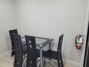 A dining area with a glass-top table and four black chairs. A fire extinguisher is mounted on the wall to the right.