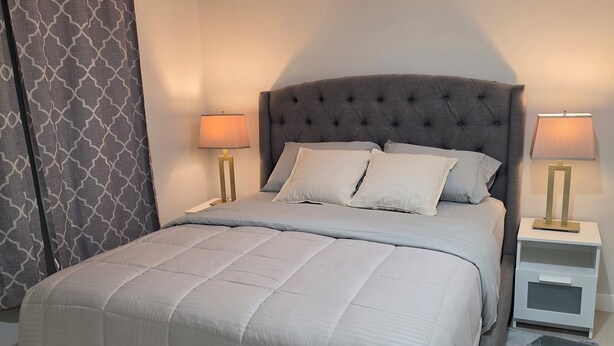 A neatly made bed with a tufted headboard, two bedside tables with lamps, and a patterned curtain. - Elegant 2 Bedroom Suite - High-End Comfort! (Calgary)