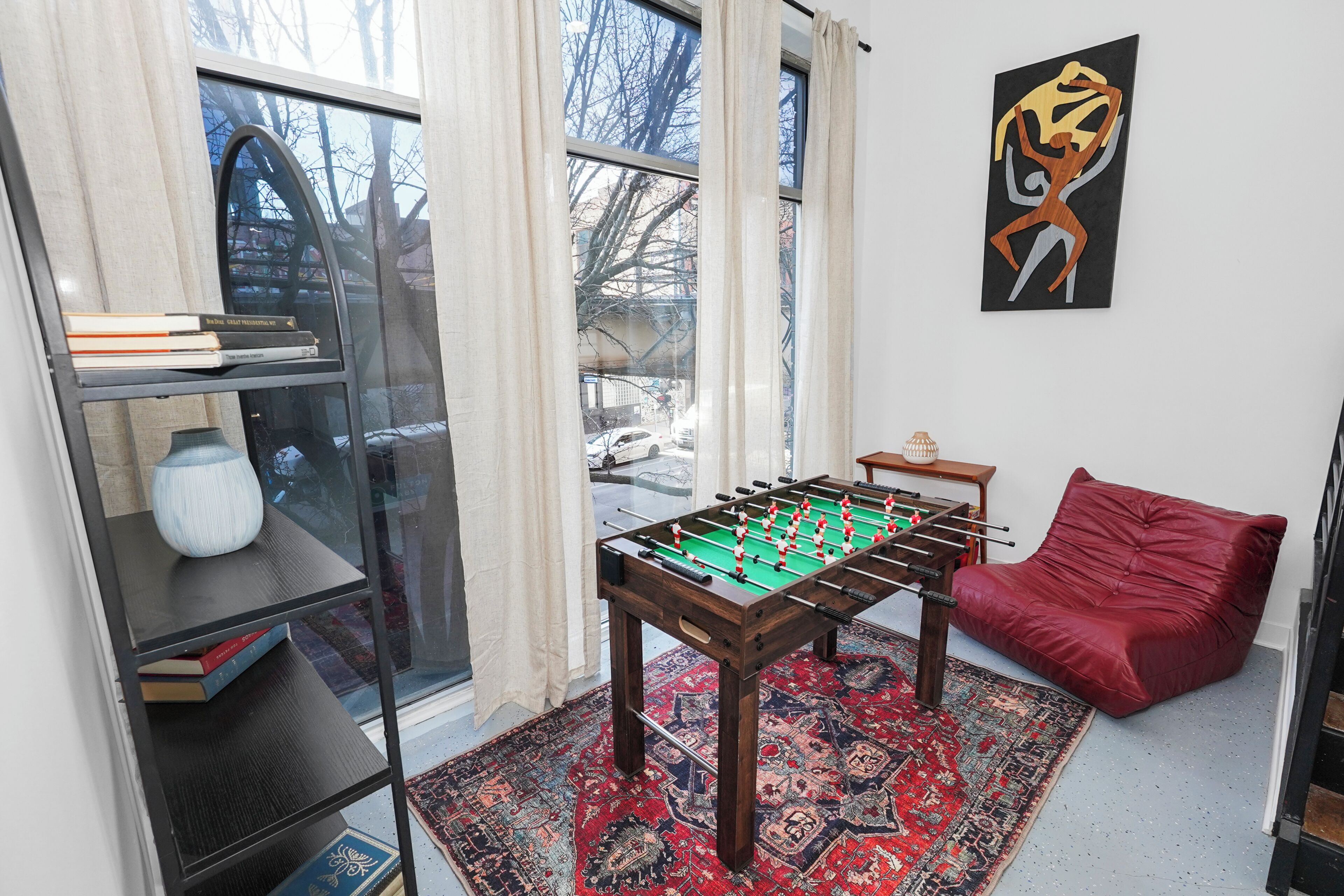 A room with a foosball table, a red sofa, a bookshelf, and a large window with a view of trees and buildings.