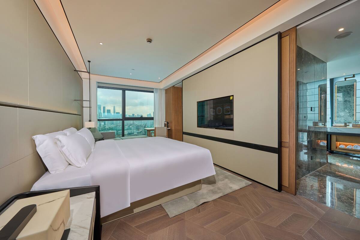 A modern hotel room with a large bed, city view, and a bathroom with a mirror and sink.