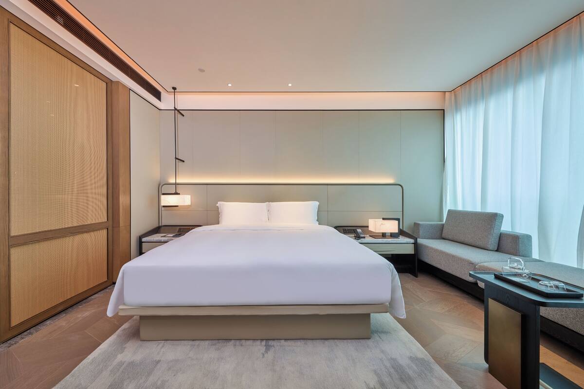 A modern hotel room with a large bed, a bedside table, a nightstand, a sofa, and a window with curtains.