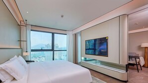 Quarto de hotel moderno com uma cama grande, uma televisão na parede e vista para a paisagem urbana pela janela.