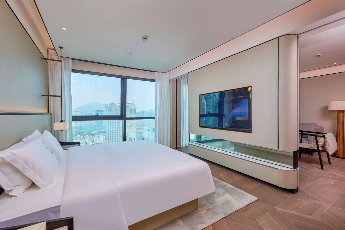 A modern hotel room with a large bed, a TV mounted on the wall, and a view of the cityscape through the window.