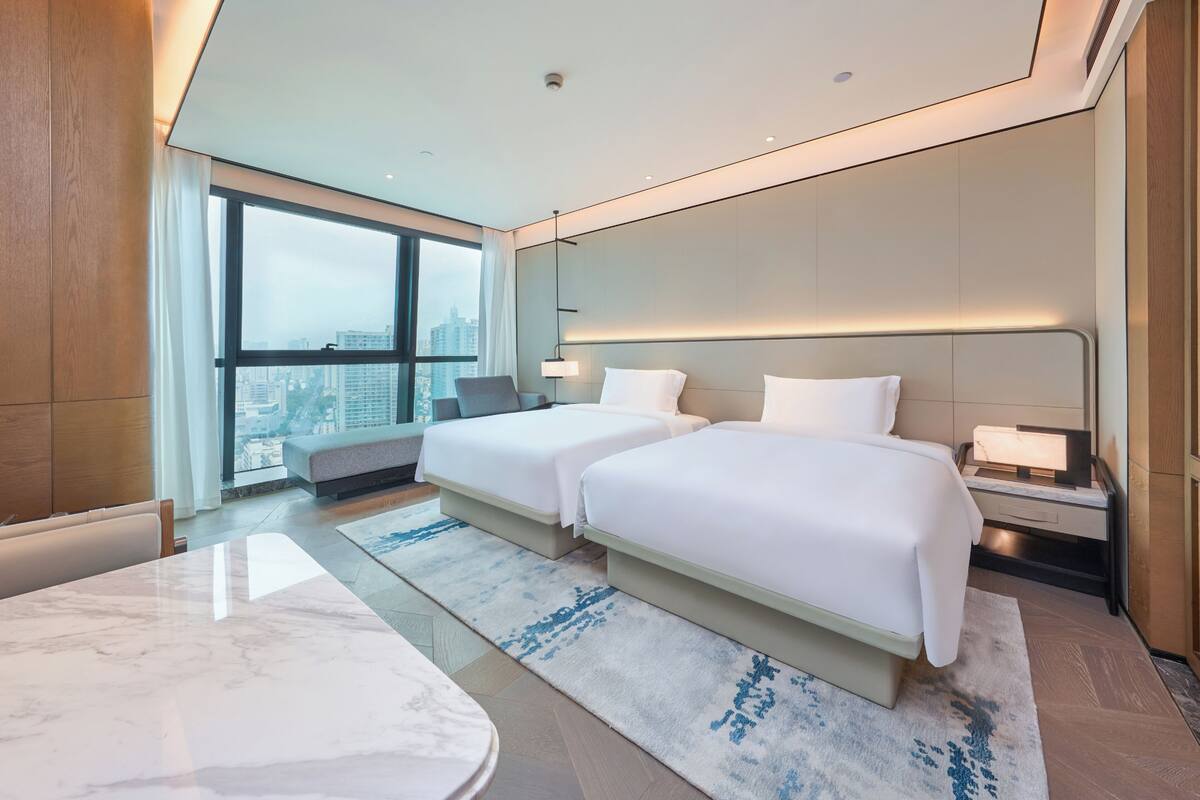 A modern hotel room with two beds, a dining table, and a large window offering a city view.