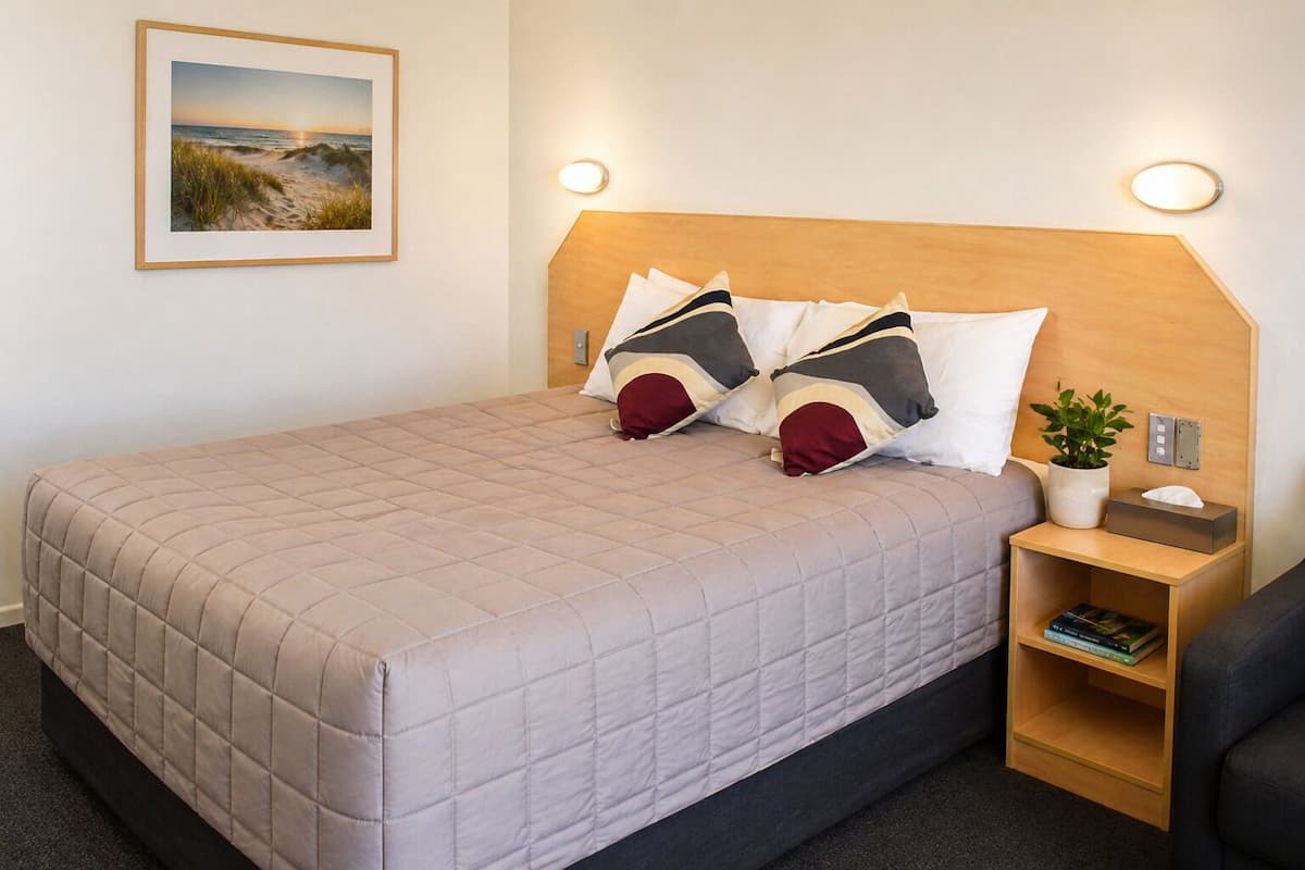 A neatly made bed with a quilted cover, a headboard with integrated lighting, a bedside table with a plant and a tissue box, and a framed picture on the wall.