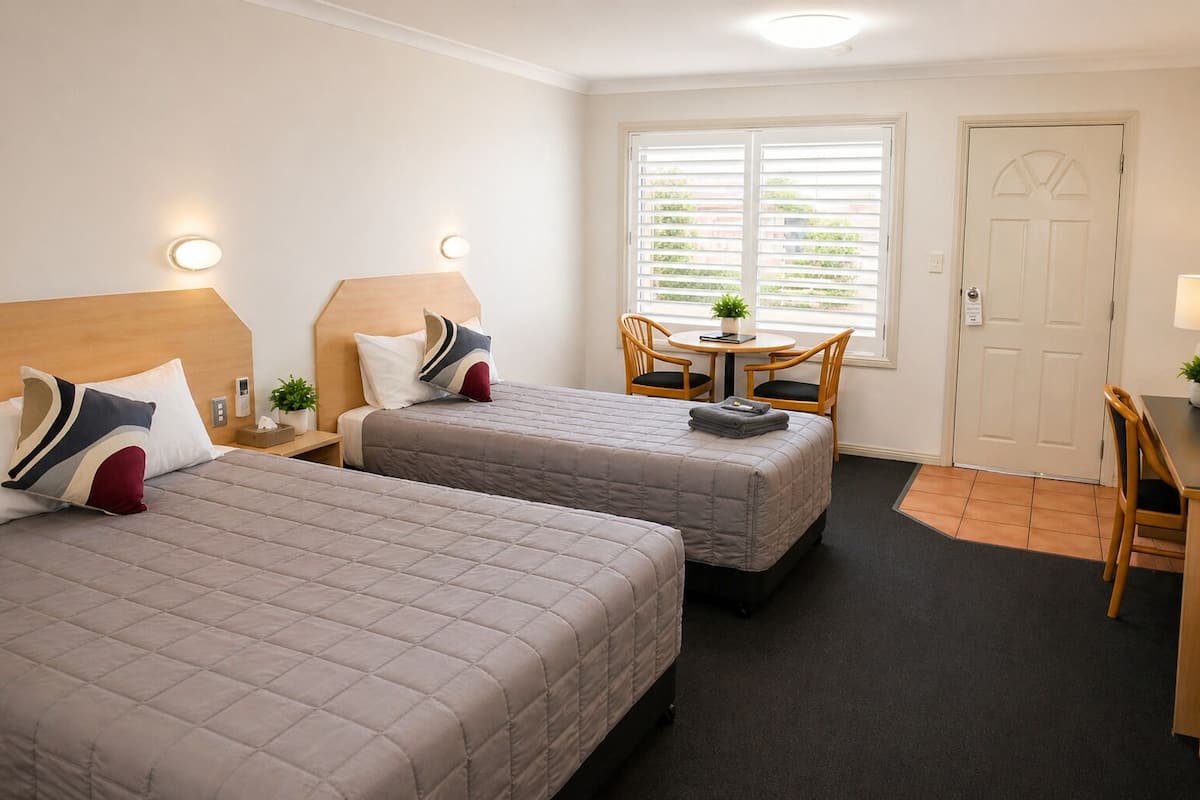 A hotel room with two beds, a small table with chairs, a door, and a window with blinds.