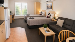 Habitación de hotel compacta con dos camas, un sofá, una mesita, un televisor, zona de cocina y una ventana con persianas.