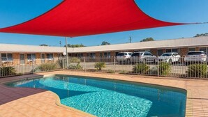 Una piscina con una vela parasol roja, área cercada y un edificio con varias ventanas al fondo.