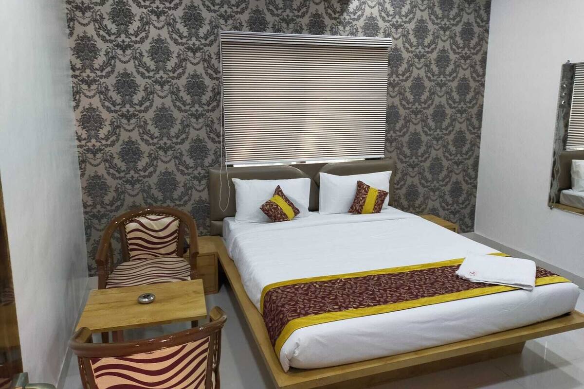 A hotel room with a bed, a wooden chair, a small table, and a window with blinds.