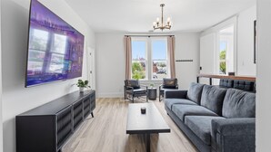 Ein modernes Wohnzimmer mit Flachbildfernseher, grauem Sofa, Couchtisch und Essbereich mit Stühlen und kleinem Tisch.