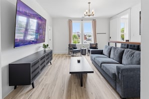 Ein modernes Wohnzimmer mit Flachbildfernseher, grauem Sofa, Couchtisch und Essbereich mit Stühlen und kleinem Tisch.