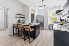 A modern kitchen with a central island, bar stools, a stainless steel refrigerator, and a window with a view. - Brand New 8 BR Double Unit House near Downtown (Indianapolis)