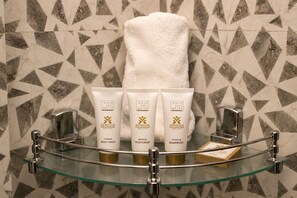A bathroom with a glass shelf holding three toiletries, a towel, and a soap bar. - Xenious Smart Suites Greater Noida (Greater Noida)