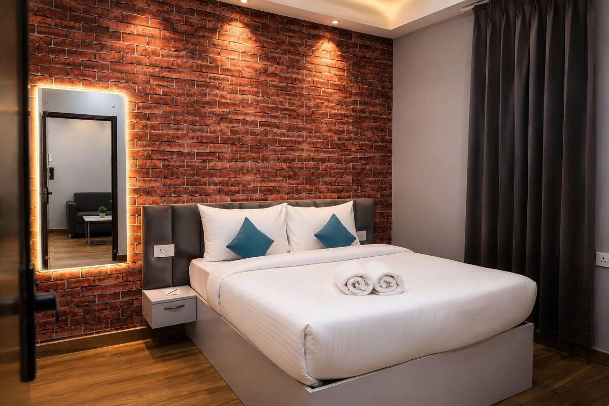 A hotel room with a brick wall, a bed with white bedding, a nightstand, a mirror, and a dark curtain.