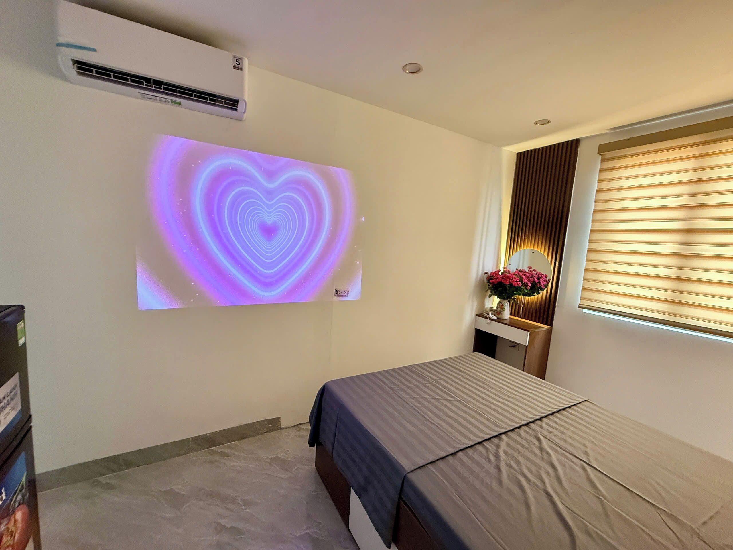 A bedroom with a bed, an air conditioning unit, a wall-mounted projector displaying a heart pattern, a bedside table with a vase of flowers, and a window with blinds.