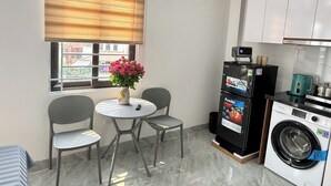 A compact kitchen with a washing machine, a table with chairs, a window with blinds, and a vase with flowers. - Era Apartment Hoang Hoa Tham (Hanoi)