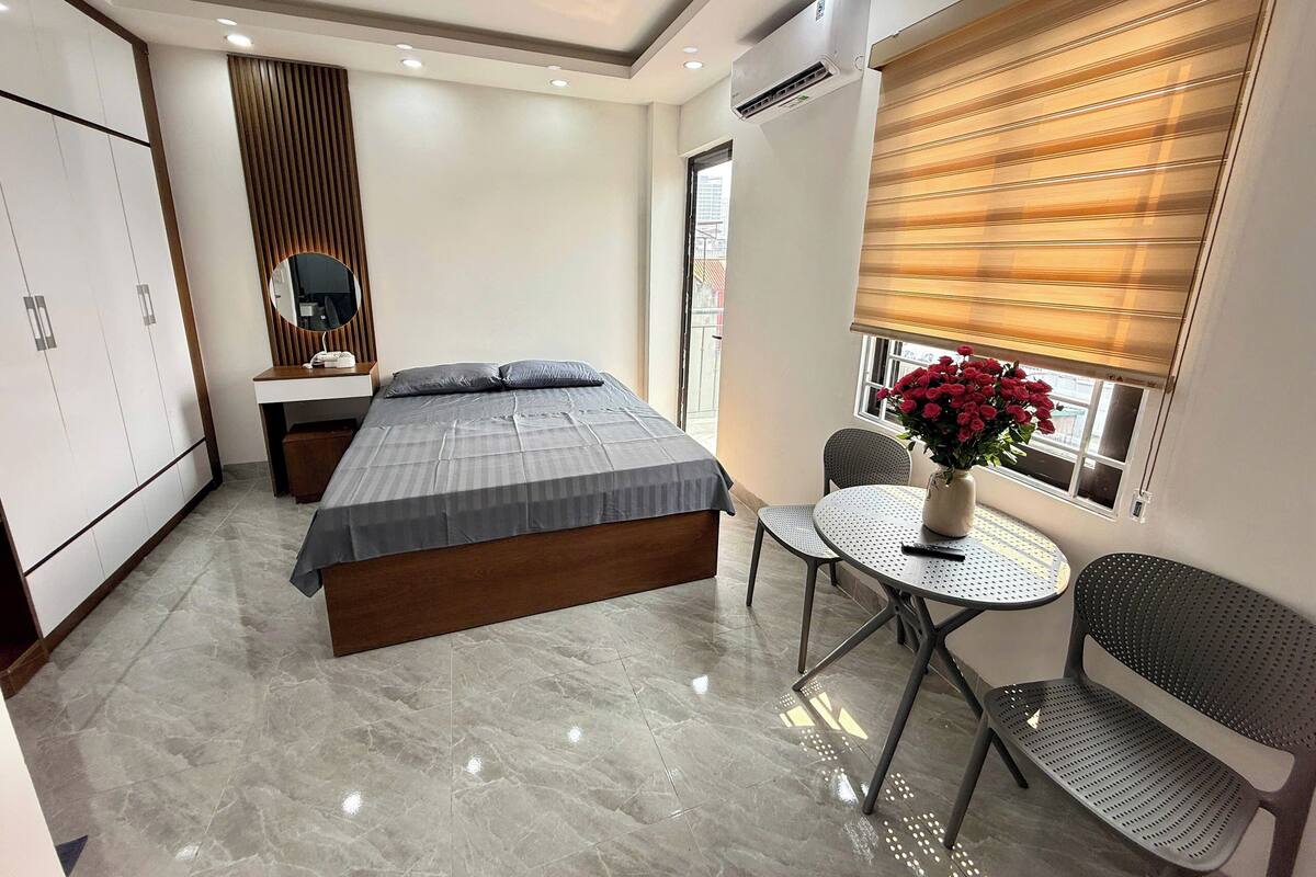 Un dormitorio con una cama, una mesita de noche, un espejo, una ventana con persianas, una pequeña mesa redonda con una silla y una planta en maceta.