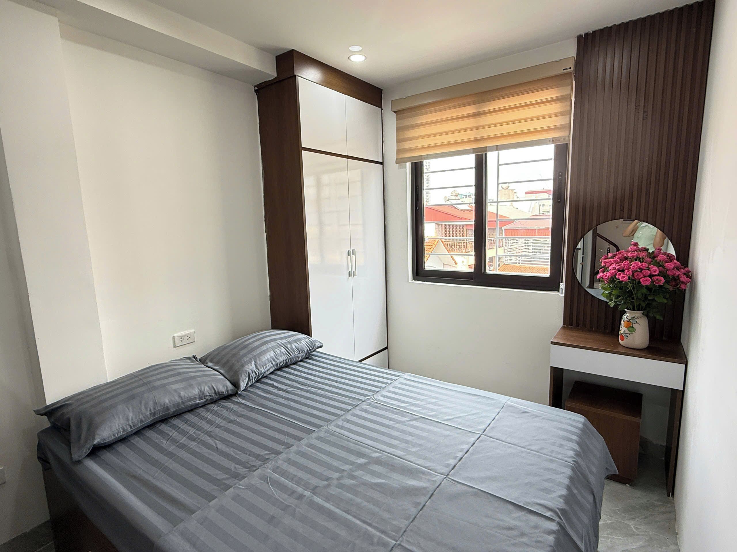 Une chambre à coucher comprenant un lit, une armoire, un bureau avec un miroir et une fenêtre donnant sur des immeubles.