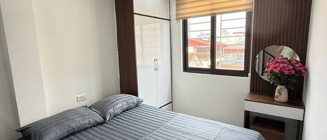 Une chambre à coucher comprenant un lit, une armoire, un bureau avec un miroir et une fenêtre donnant sur des immeubles.