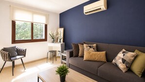 Een moderne woonkamer met een donkerblauwe accentmuur, een grijze bank met gekleurde kussens, een houten salontafel, een wit bureau met stoel, en een raam met uitzicht op groen.
