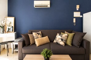 A modern living room with a dark blue wall, a grey sofa with patterned cushions, a small wooden coffee table, and a potted plant. - Mia 1-BR Apartment & Balcony in Larnaca (Larnaca)
