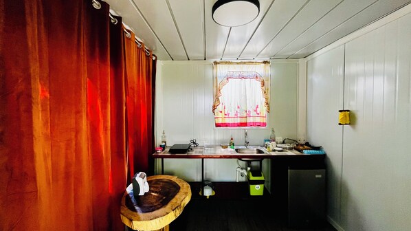Une cuisine compacte avec un plan de travail en bois, un évier, une fenêtre ornée d’un rideau à motifs floraux et un petit tabouret.