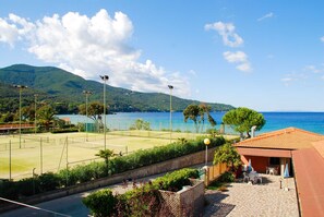 A coastal view with a tennis court, a building with outdoor seating, and mountains in the background. - Elbamar Procchio - Elbamar Procchio - TRILO 6 Cottage (Marciana)
