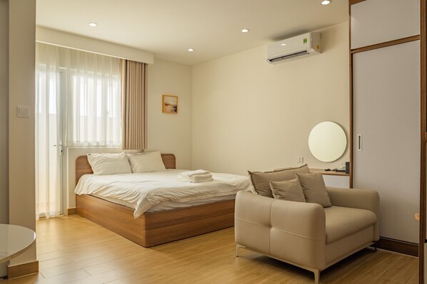 A modern hotel room with a wooden bed, a beige sofa, a round mirror, and a window with sheer curtains. - CHOWA HOME Thao Dien by DNM Hospitality (Ho Chi Minh City)