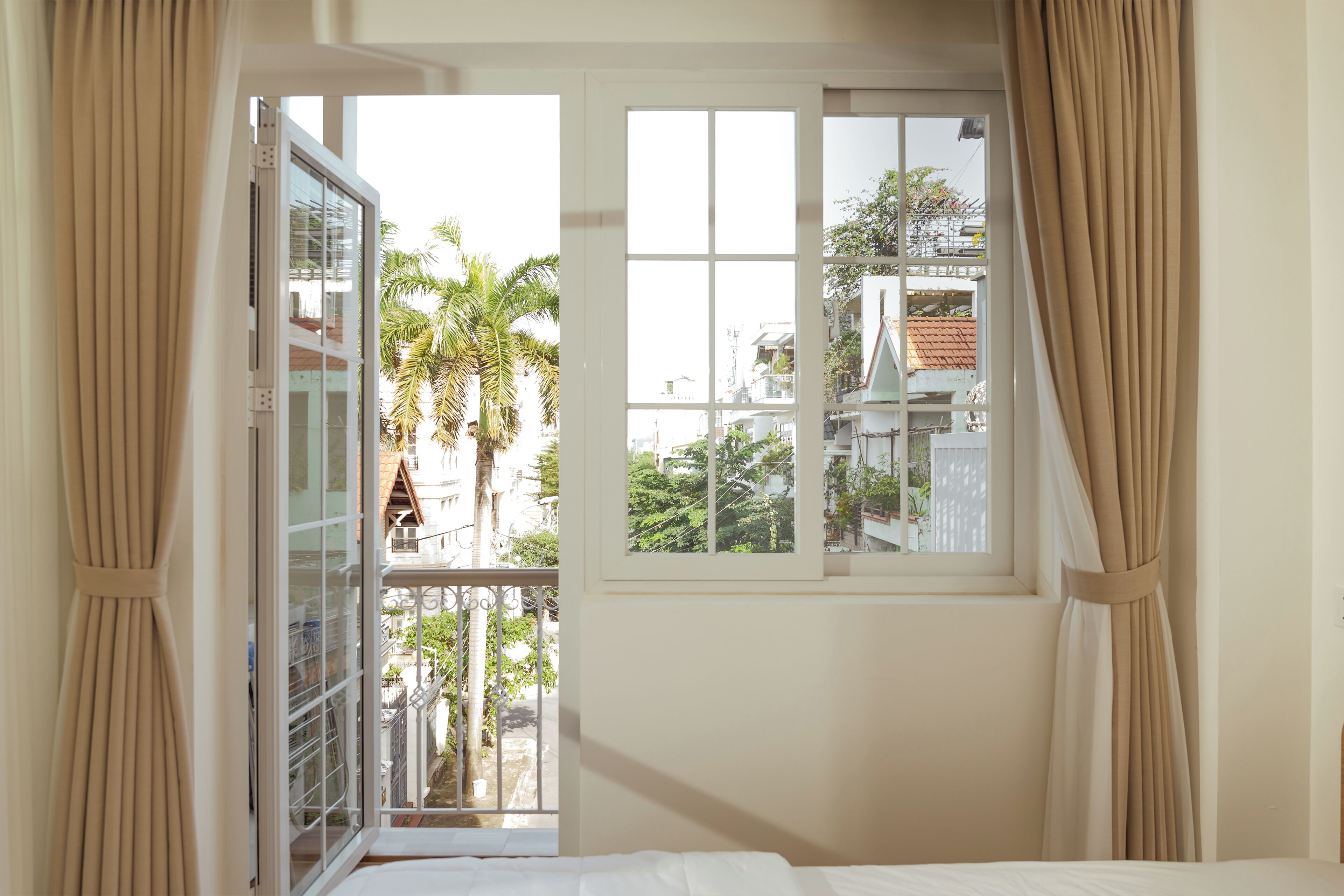 Quarto com cama, janela com cortinas e vista para edifícios e palmeiras lá fora.