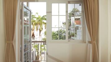Quarto com cama, janela com cortinas e vista para edifícios e palmeiras lá fora.