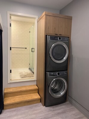 Un lavadero con lavadora y secadora de carga frontal, escalones de madera y una zona de ducha con azulejos.