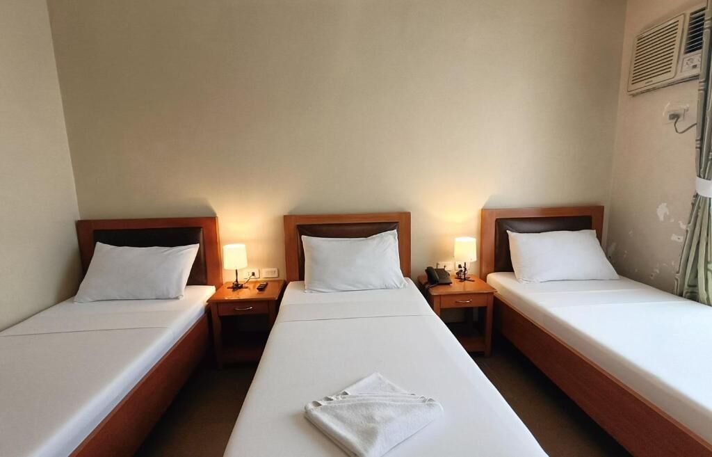 Una habitación de hotel con tres camas individuales, cada una con cabecero de madera y ropa de cama blanca. Hay mesitas de noche con lámparas y un teléfono en el lado derecho de la habitación.