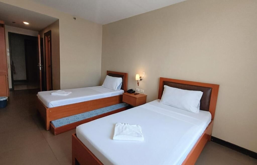 A hotel room with two single beds, a nightstand with a lamp, and a visible door leading to another room.
