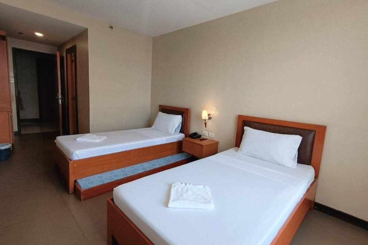 Habitación de hotel con dos camas individuales, una mesita de noche con lámpara y una puerta visible que da a otra habitación.