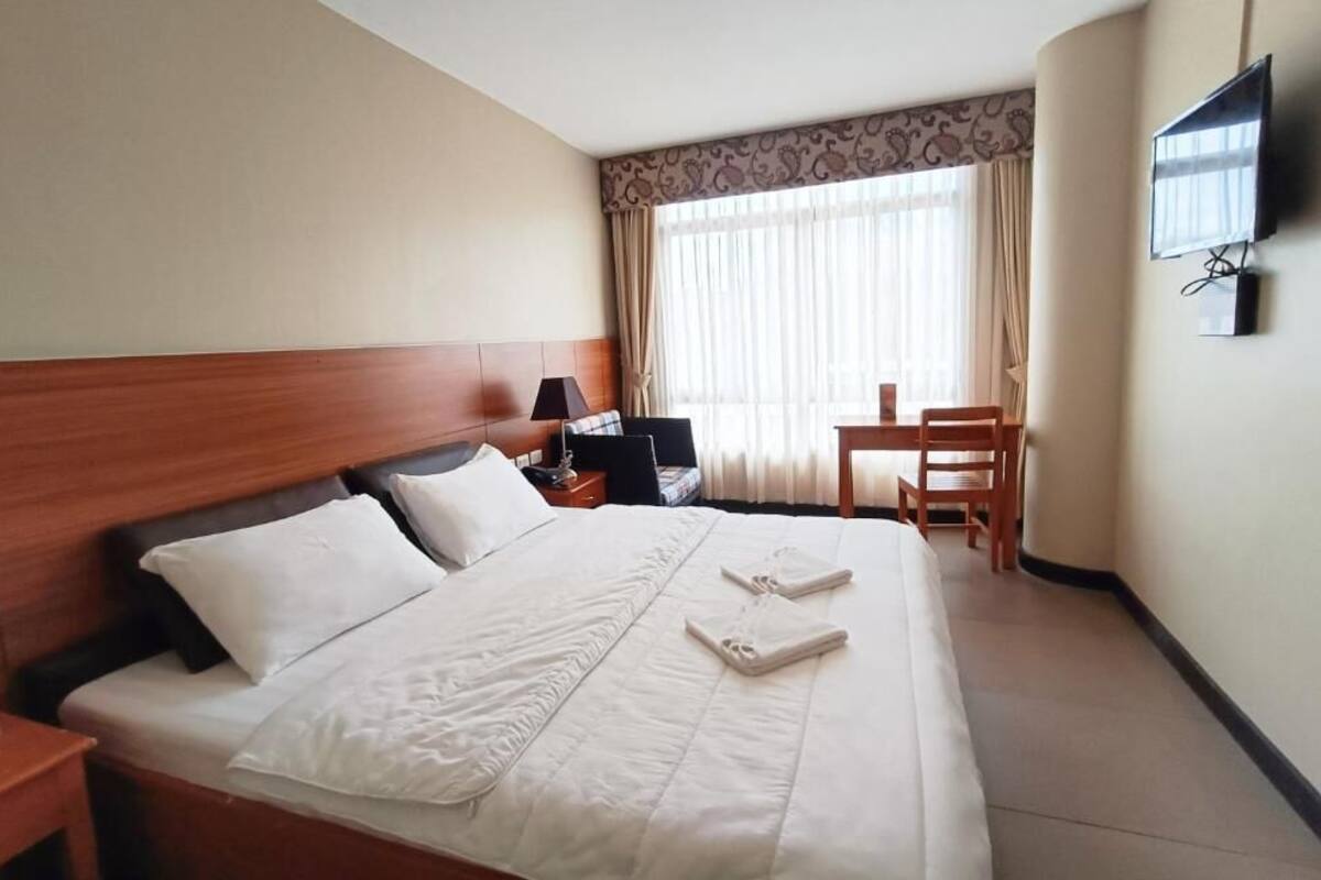 Habitación de hotel con cama, mesita de noche, lámpara, televisor y escritorio con sillas.