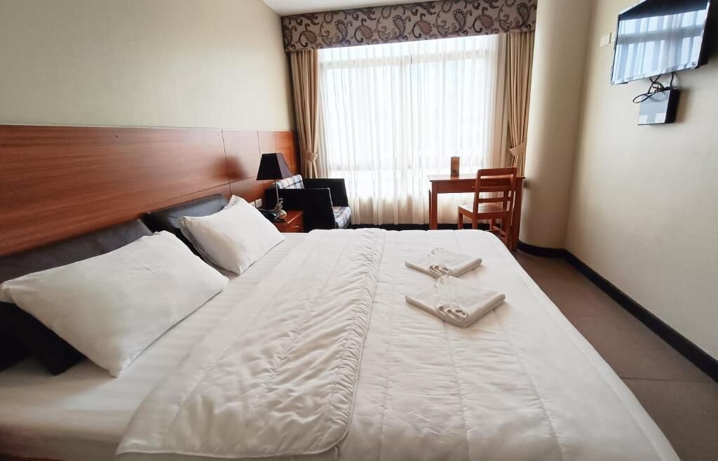 A hotel room with a large bed, a desk, a chair, a TV, and a window with curtains.