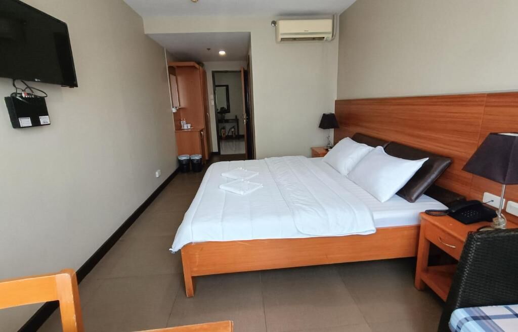 A hotel room with a wooden bed, a flat-screen TV mounted on the wall, a bedside table with a lamp, and a chair with a checkered cushion.