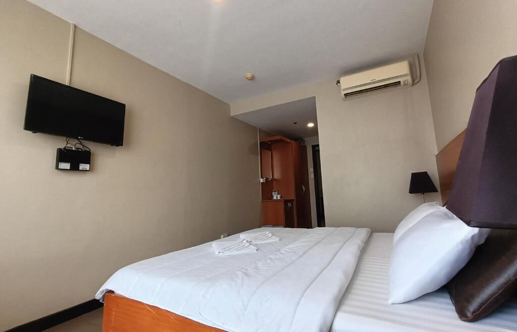 A hotel room with a bed, a flat-screen TV mounted on the wall, a wooden wardrobe, and an air conditioning unit.