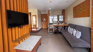 Una sala de estar con televisor de pantalla plana, un sofá gris con cojines estampados, una pared de acento de madera y una cocina al fondo.