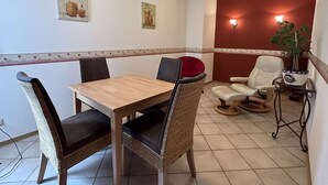 A dining area with a wooden table and wicker chairs, a red cushioned chair, a white chair, and a small table with a plant. - Cozy vacation apartment with balcony - 2 bedrooms with 2 private bathrooms (Erden)