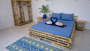 Una cama con colcha azul, dos almohadas con forma de corazón, una planta en maceta y un cabecero de bambú tejido.