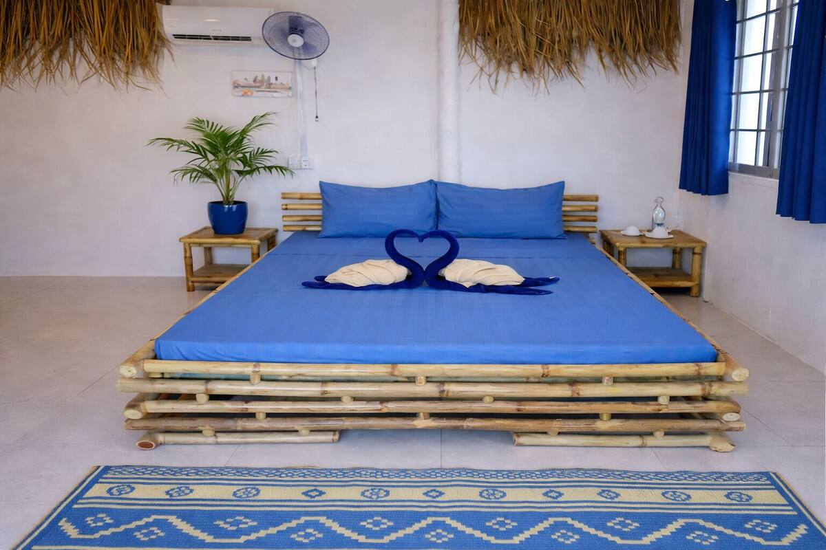 A bed with a blue bedspread, two blue pillows, a heart-shaped towel arrangement, a potted plant, and a blue and white patterned rug.