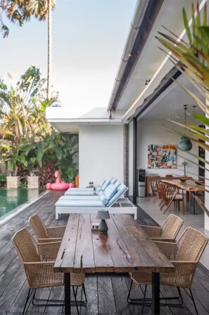 A wooden deck with a dining table and chairs, a pool, lounge chairs, and a modern kitchen visible through an open door. - “Exclusive 3-Bedroom Villa – Perfect for Shared Living with Family” (Tibubeneng)