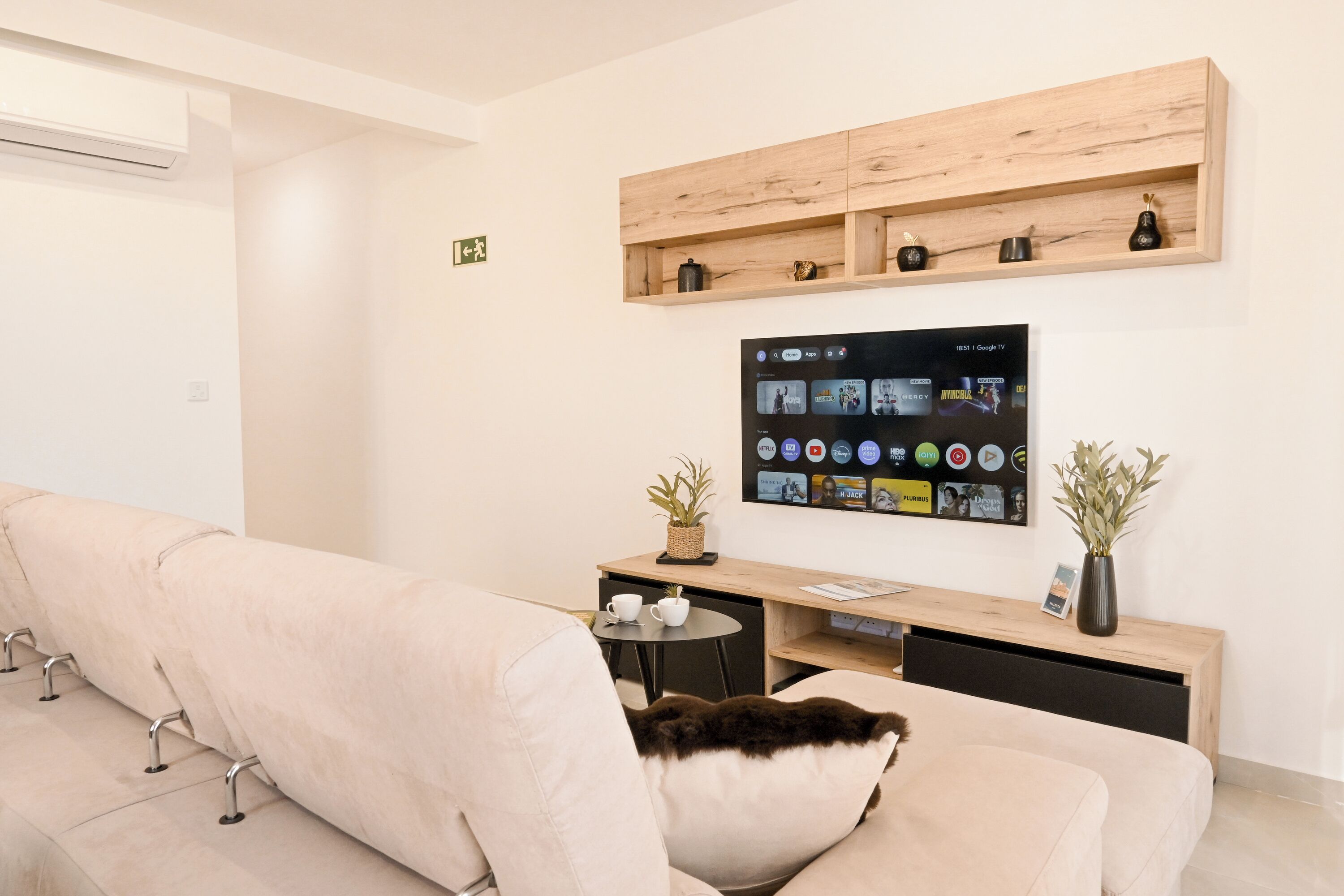 Un salon moderne avec un canapé, une table basse, un téléviseur fixé au mur et une étagère en bois au-dessus.