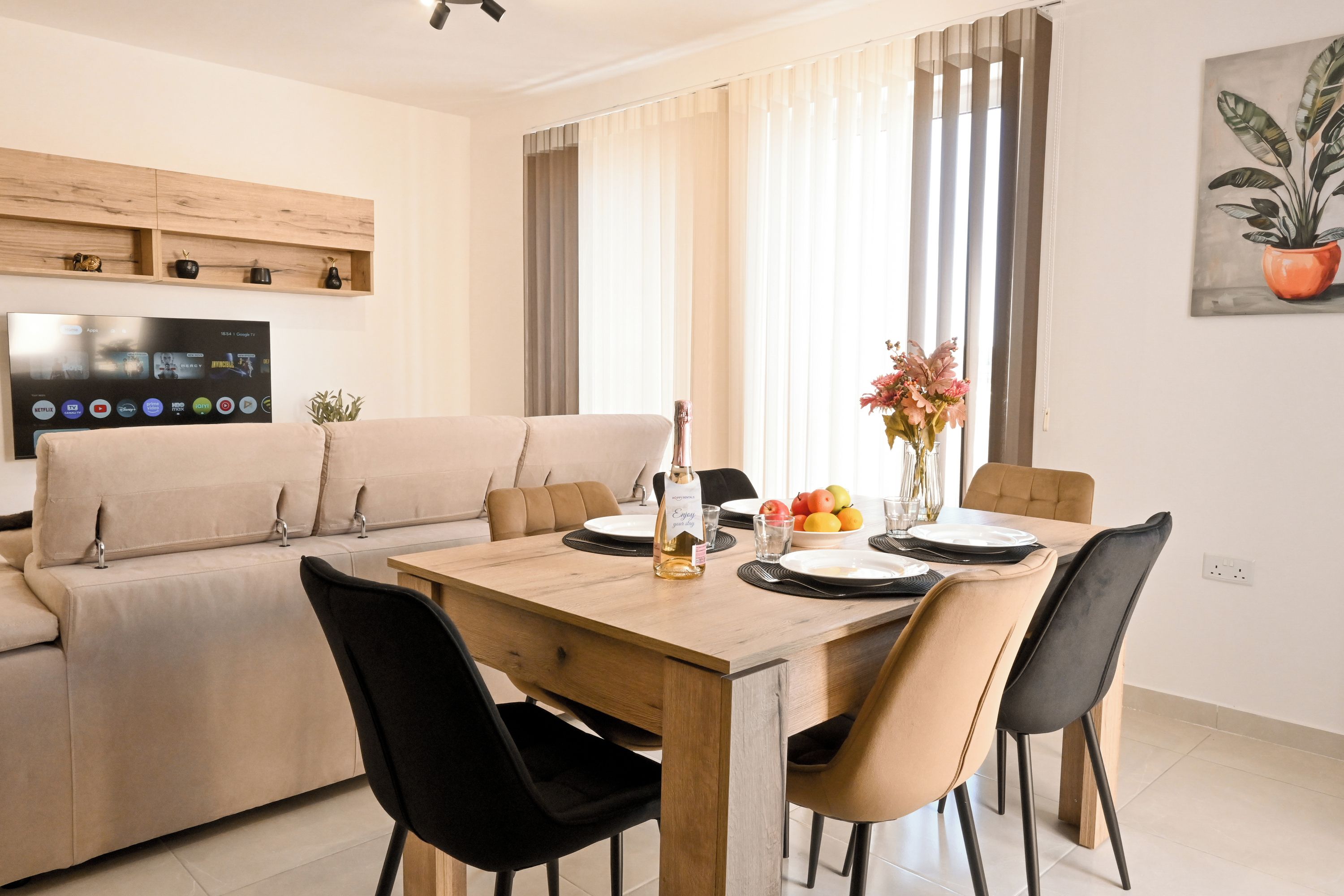 A modern living room with a sofa, dining table, chairs, a TV mounted on the wall, and a potted plant.