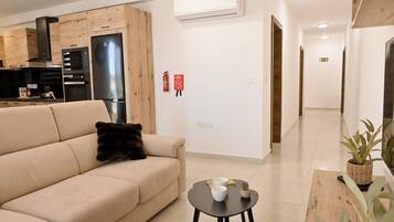 A modern living room with a sofa, a small table with decorative items, a kitchen area with appliances, and a hallway with doors.
