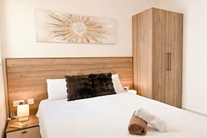 A hotel room with a wooden headboard, a white bedspread, a black fluffy pillow, a white pillow, a wooden wardrobe, and a wall-mounted abstract art piece. - Viviani Court 2bd Modern Apt Xewkija Gozo (Xewkija)