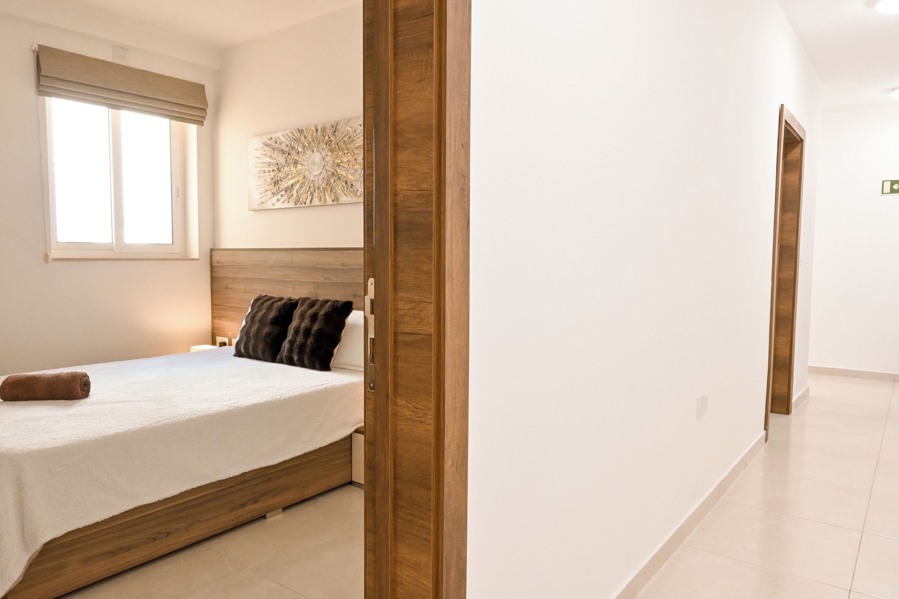 A bedroom with a wooden headboard, a bed with a white bedspread, a window with blinds, and a wall-mounted decorative piece.