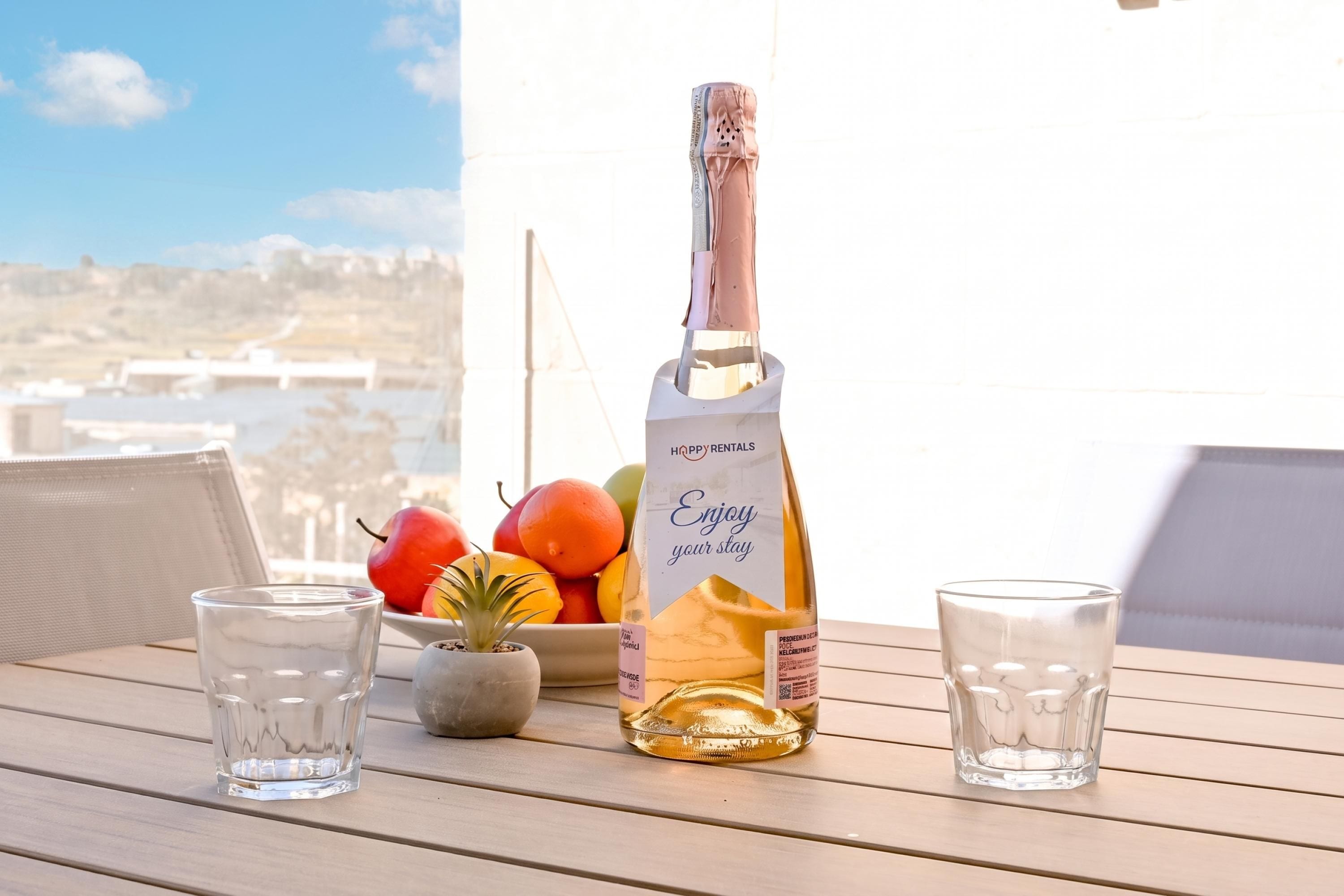 A bottle of alcohol, two glasses, and a bowl of fruit on a wooden table with a view of a body of water and buildings in the background.